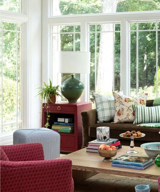 conservatory living room with white trim, floor-to-ceiling windows, brown sofa with colorful patterned cushions and red armchair and red side table with green lamp