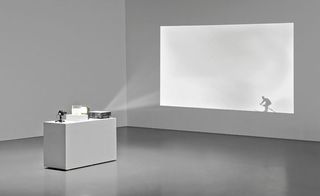 A room with white walls and white rectangular box in the middle with a projector placed on it and projecting on the wall a man snowboarding