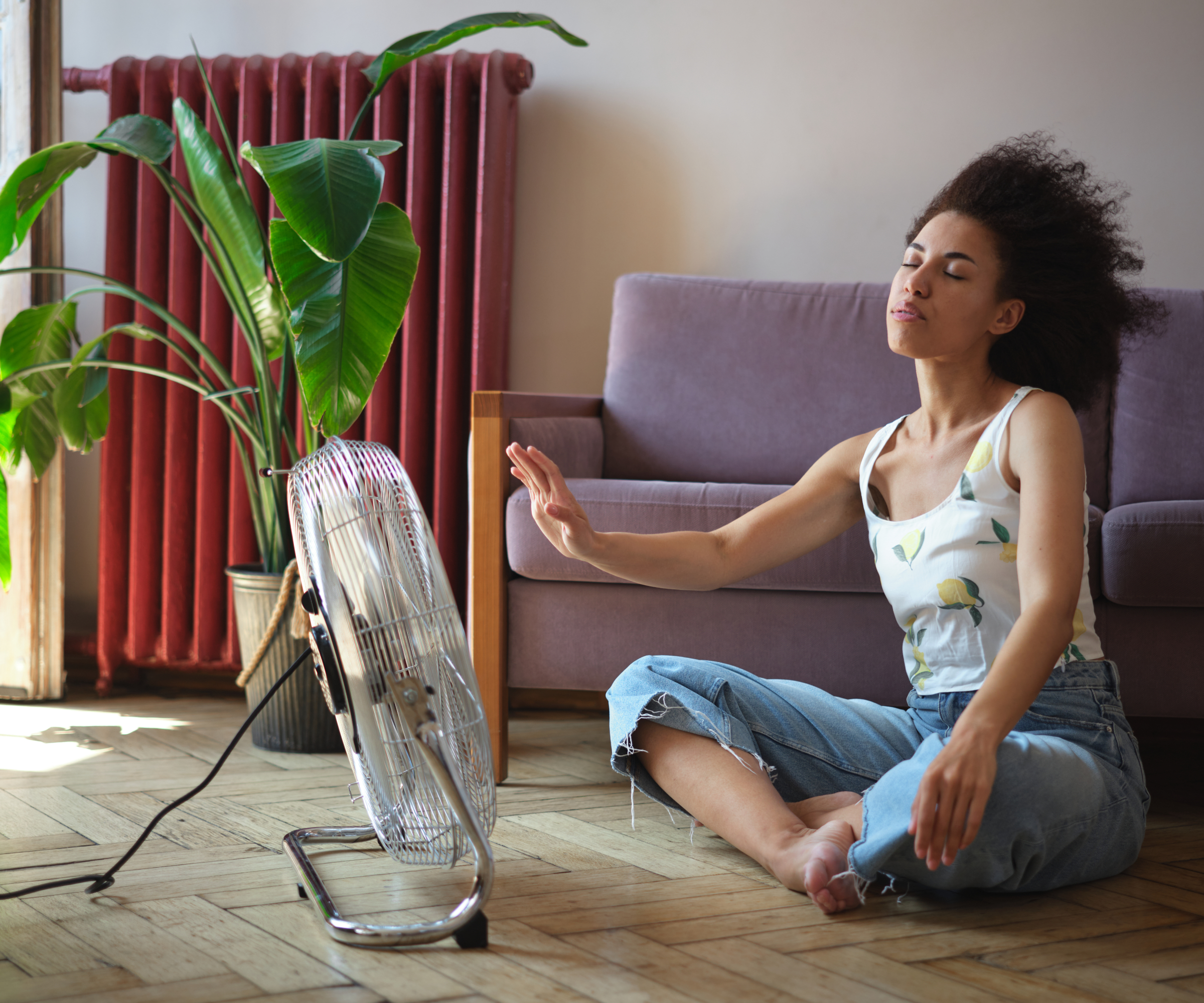 Happy contented African American curly woman using electric fan at home enjoying fresh air on hot summer day