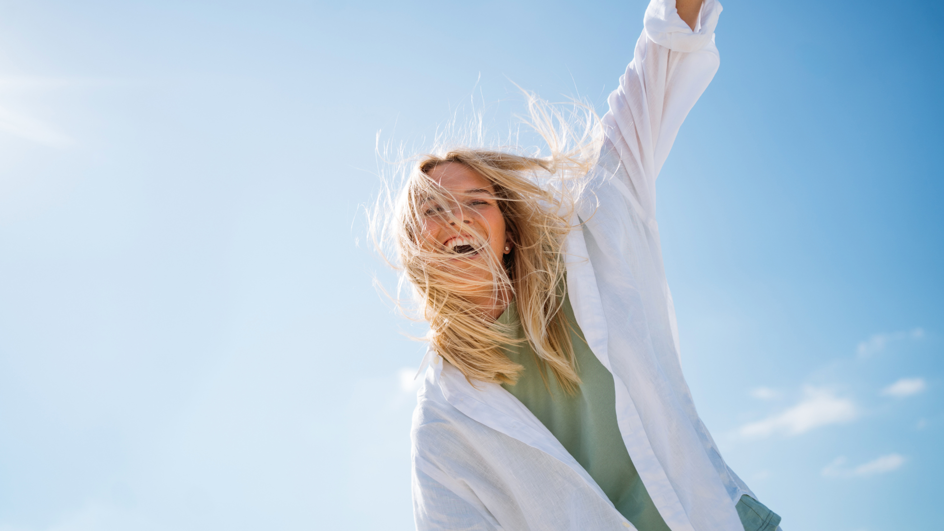 A woman smiles and stretches one arm into the air as she stands outside in the sun.