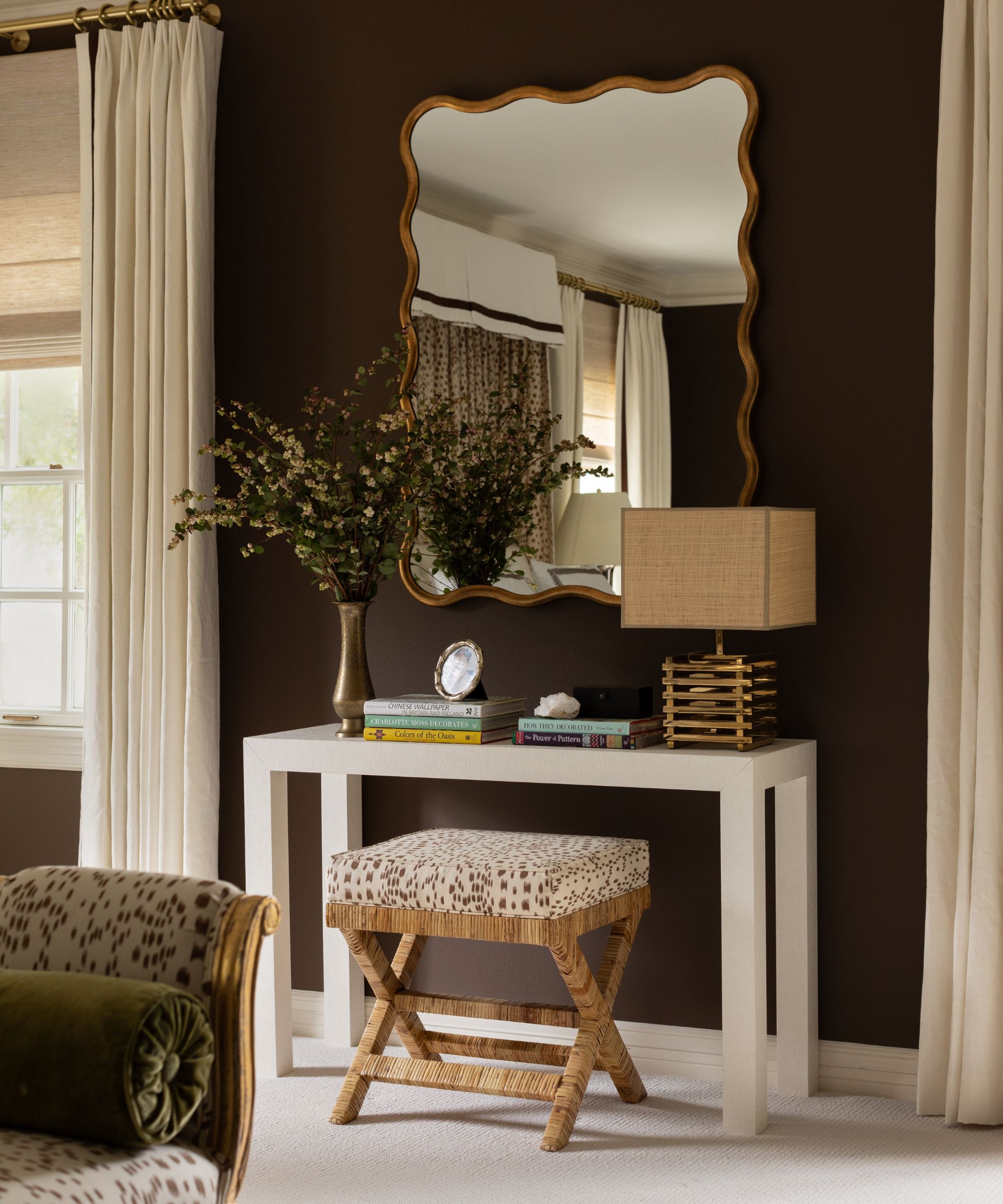 A bedroom with chocolate brown walls, white trim, a cream carpet, cream curtains, a wavy mirror above a white vanity with a lamp on it, and a vase of flowers.