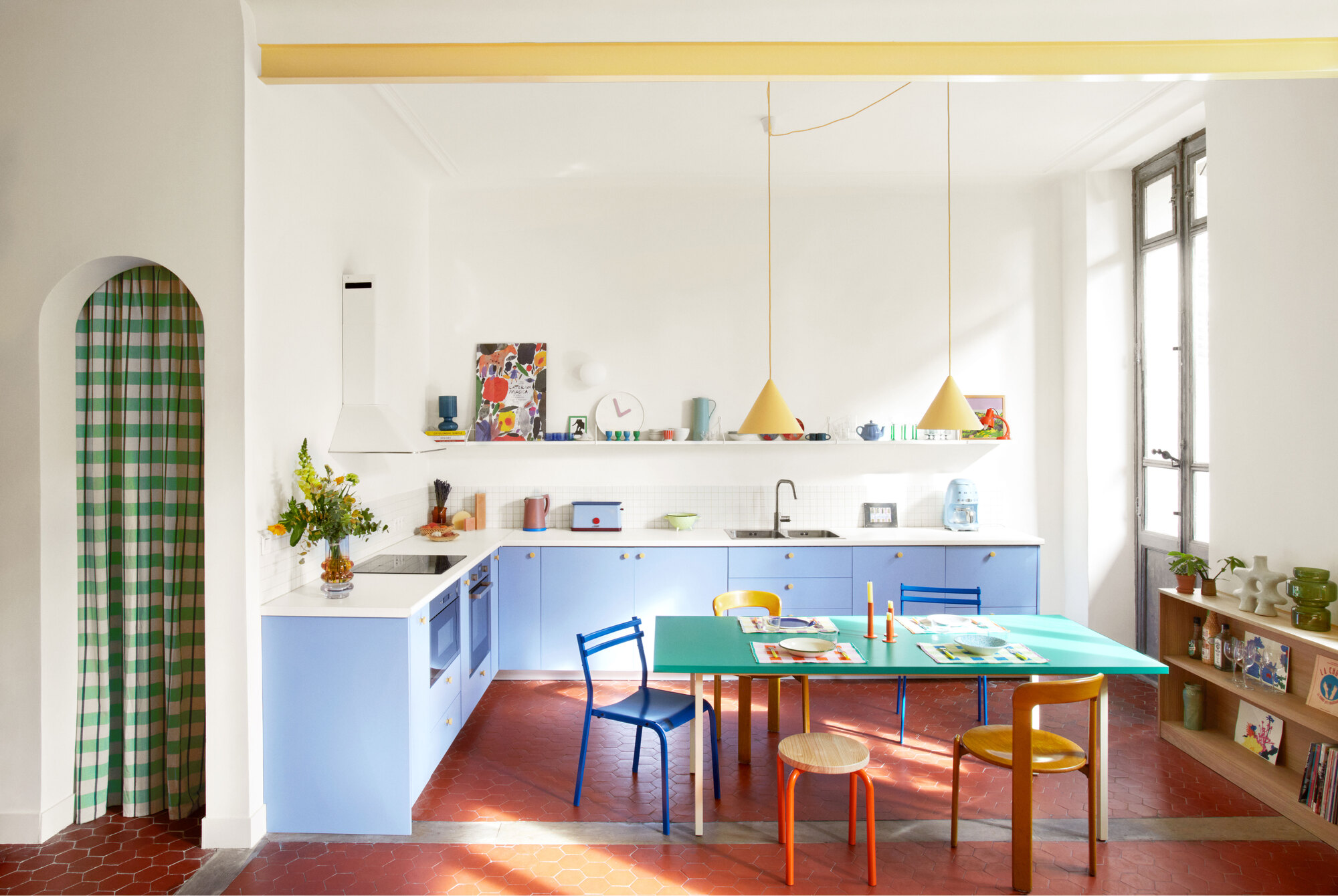 colorful kitchen with arched doorway with checked curtain, light blue cabinetry, white counters, open shelving with colorful decor, red floors, green dining table with colorful chairs, and timber console styled with books