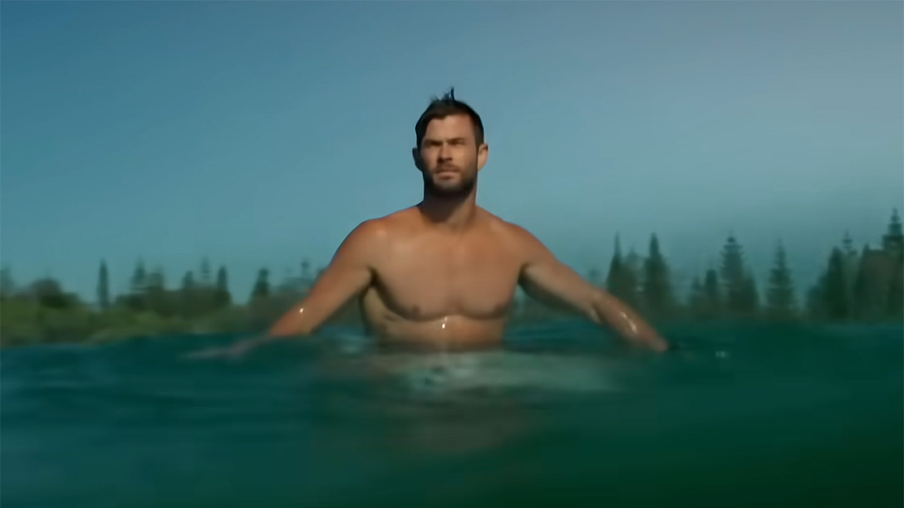 Chris Hemsworth screenshot of him surfing shirtless in bright blue water from video footage.