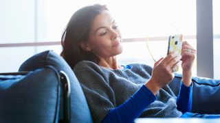 Woman looking at phone, sitting on sofa at home with sunlight coming through window behind her
