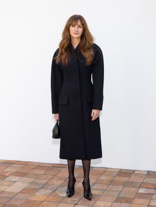 Julia Roberts stands in front of a white wall at the Jacquemus show wearing a black coat dress, black tights, and black shoes