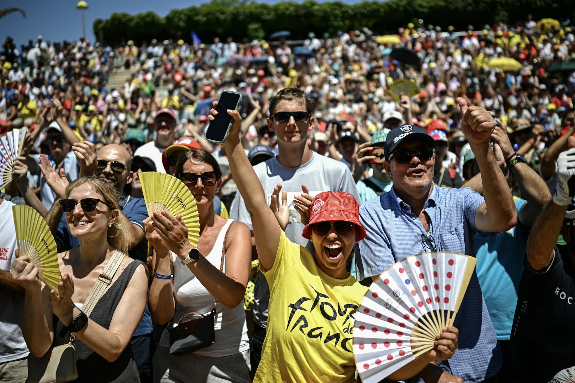 A woman in a bucket had and Tour de France yellow shirt holds a polka dot fan in front of a large crowd of Tour de France specators