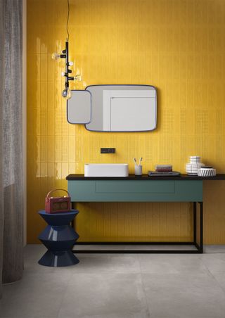 yellow tiled bathroom with green vanity, gray floor tiles, blue trim mirror, bulb pendant light, accessories, blue stool, radio