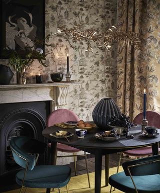 Modern dark and moody dining room with statement leaf lighting, chinoiserie design wallpaper, and assorted velvet chairs.