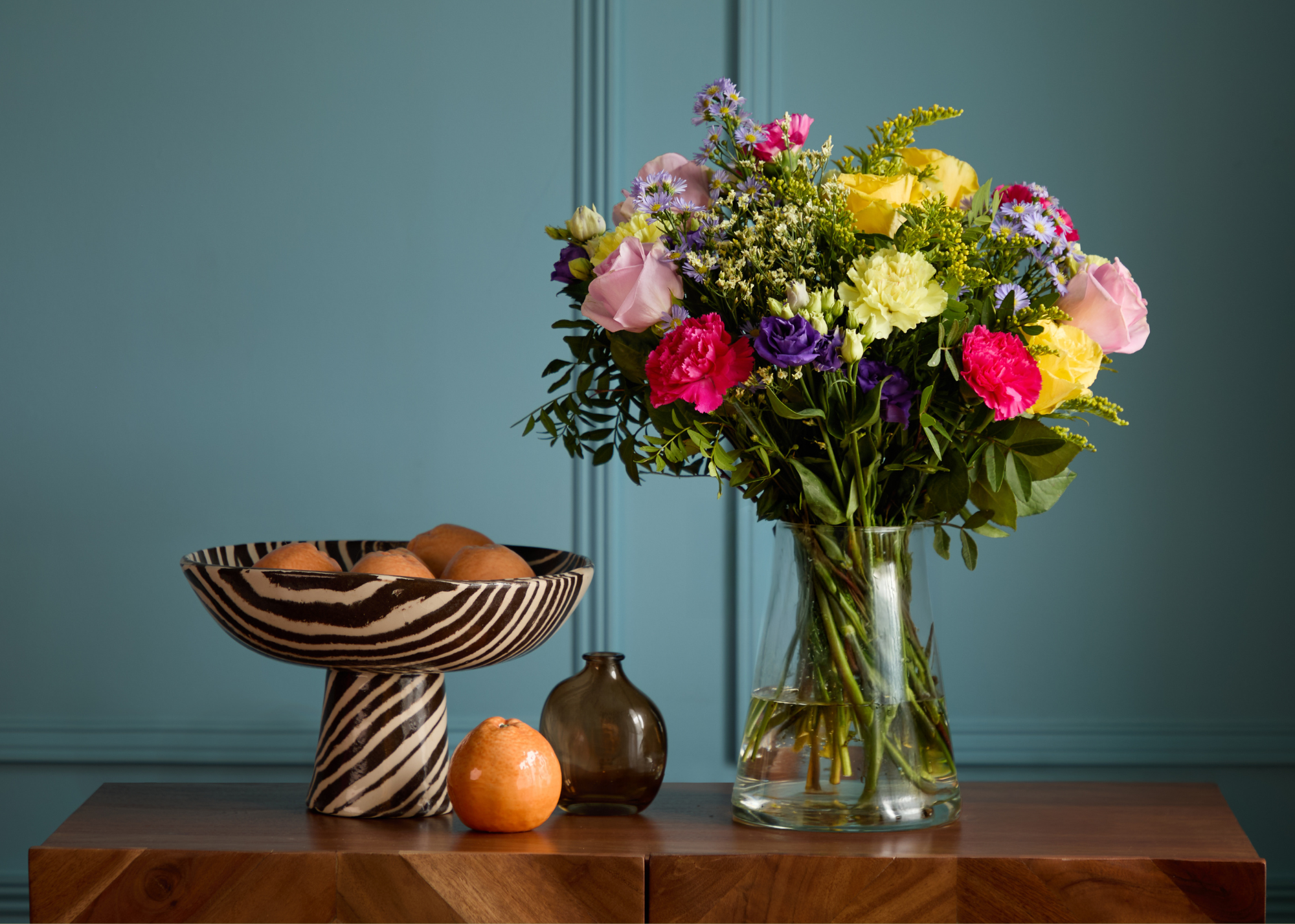 A wood sideboard with a glass vase of colorful flowers, a footed marbled bowl or oranges, and an amber vase in a blue room