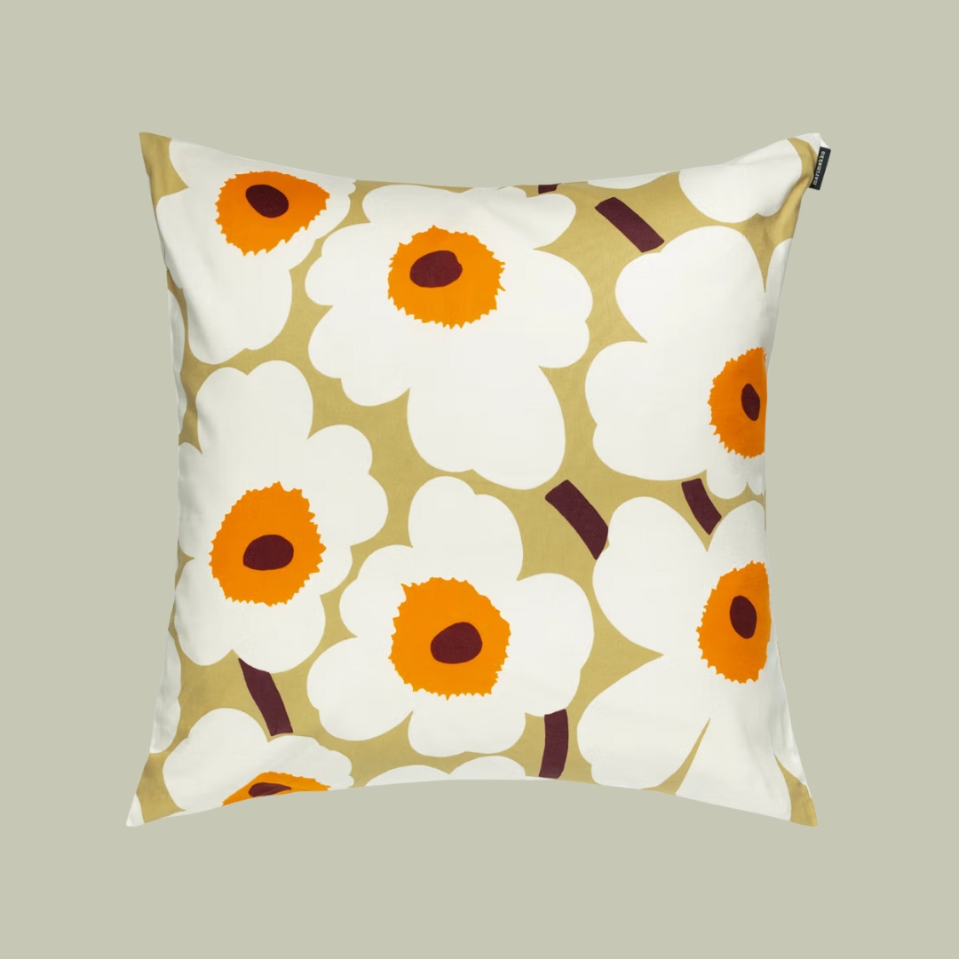 A square cushion with white flowers with a bright orange heart sits against a pale green background.