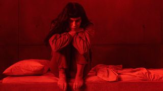 A red still of Milena Smit sitting on a bed in 