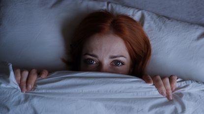 Woman hides beneath her bedcovers looking frightened.