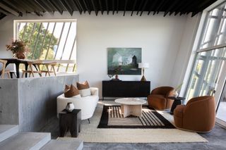 A modern living room with neutral decor including a white sofa, burnt orange chairs and a natural woven rug., juxtaposed with cement architectural features