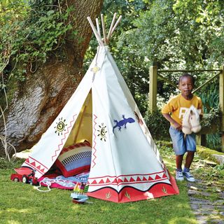 Hideout Wigwam white teepee tent decorated with red pattern, cacti, lizards and suns on it. Tent is in a garden with a little boy riding a toy horse next to it