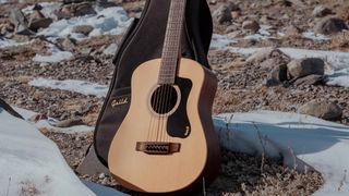 The Guild Travel Spruce acoustic is photographed on the side of a snow-covered hill-top, leaning against its gig-bag.