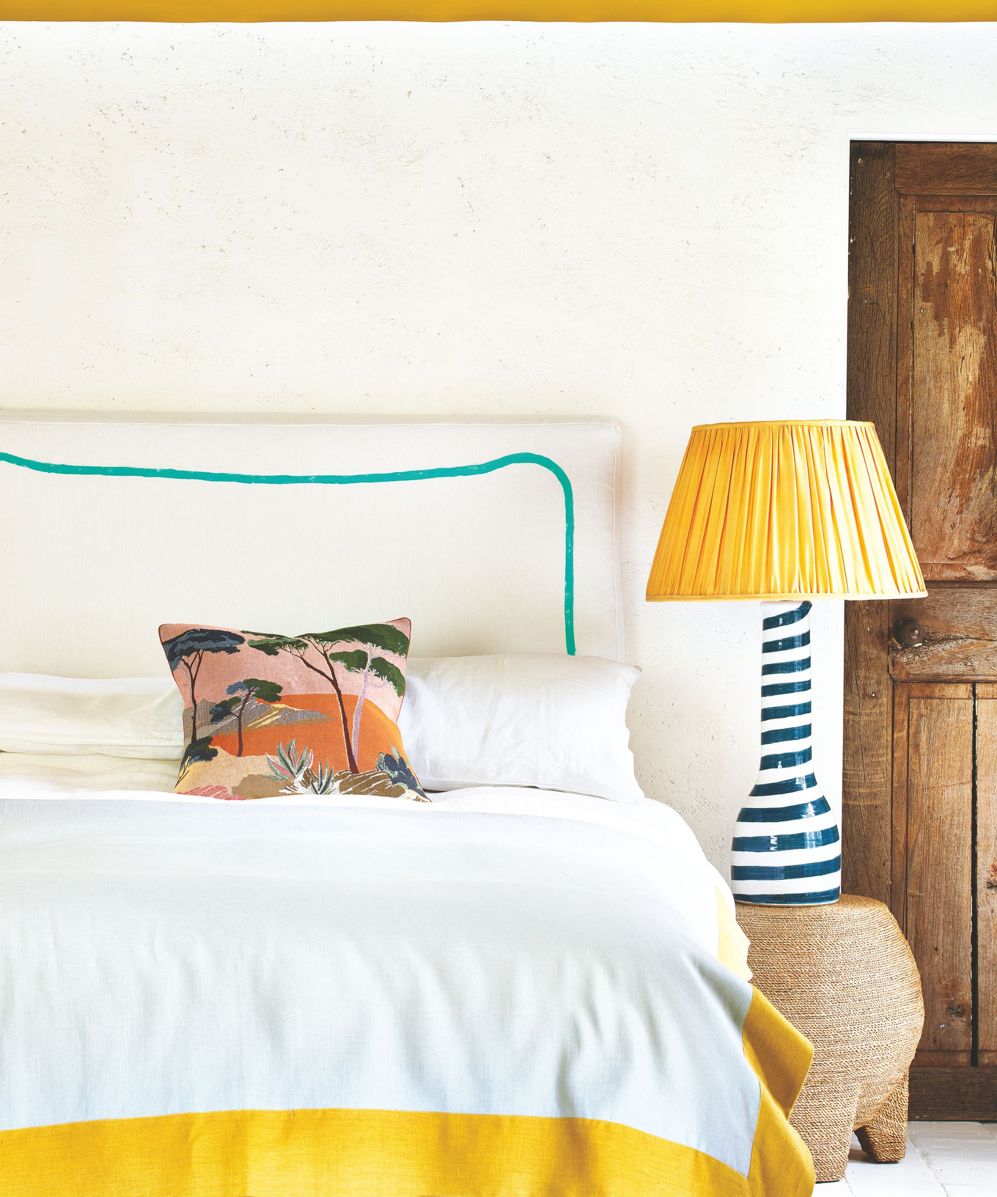 A yellow and white bedroom with a white bed with blue stripe, white pillows, decorative cushion, and a striped blue and white lamp on a rattan table, with a yellow lampshade beside a wooden door.