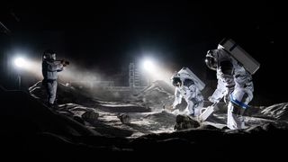 ESA astronauts Matthias Maurer and Aidan Cowley work on the simulated landscape of the moon, with stage lights recreating the lighting on the lunar surface. Another photographer dressed in protective gear stands to the left.
