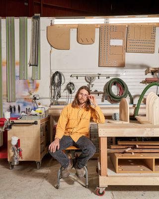 A young man dressed in a yellow workwear shirt with silver buttons, black trousers, and brown shoes sits in a wood workshop covered in utensils and artificially lit by neon lights.