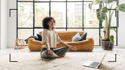 Woman meditating at home using one of the best meditation apps on her laptop, with large open window behind her 