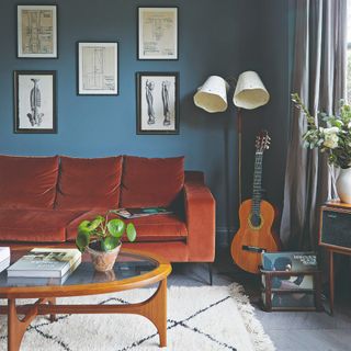 A blue-painted living room with mid-century modern furniture like a wood and glass coffee table and a rust-coloured velvet sofa