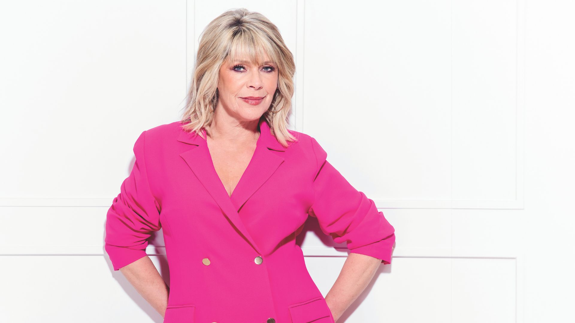 Ruth Langsford wears a hot pink suit and stands against a white panelled wall, looking glamorous
