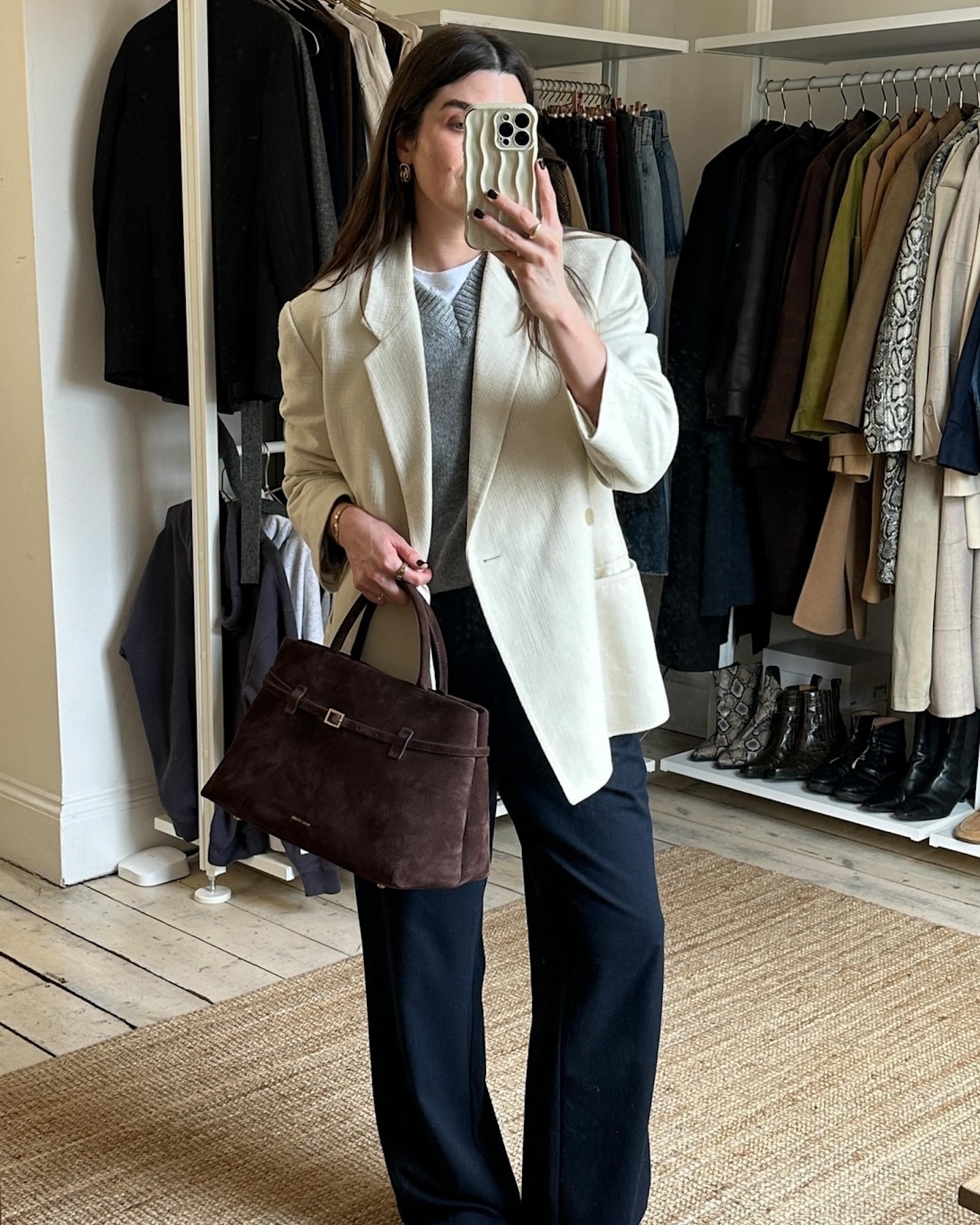 British style influencer Anna Howard poses for a mirror selfie wearing an ivory blazer, gray v-neck sweater, brown suede tote bag, and wide-leg jeans.