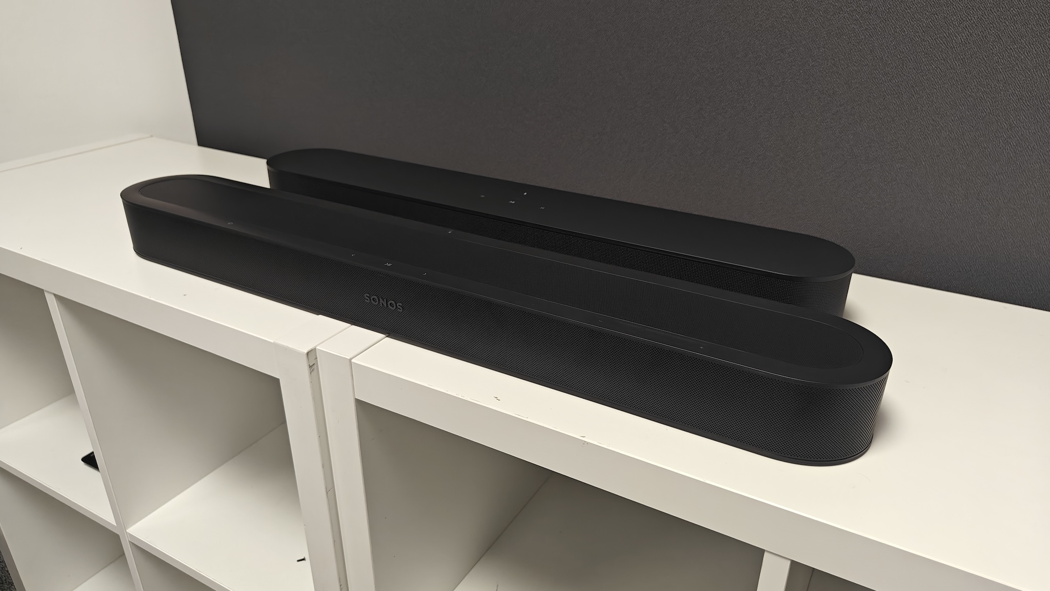 A black Sonos Beam Ultra and Sonos Beam (Gen 2) side by side on a white TV cabinet, shot at a 3/4 angle.