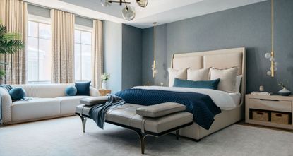A bedroom with a blue and cream color scheme, a bench at the end of the bed, and a sofa in the window