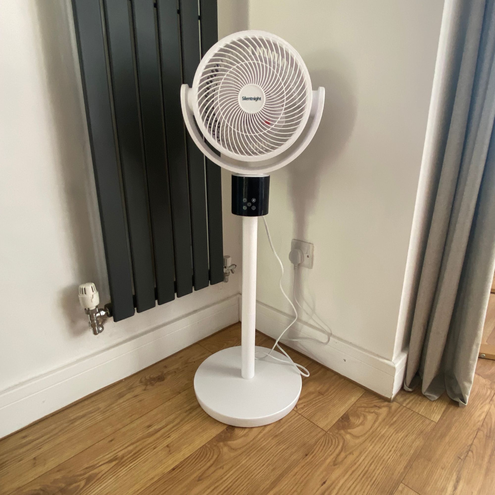 Silentnight AirMax Pro360 pedestal fan in pedestal mode on a wooden floor in front of a grey vertical radiator and a white wall