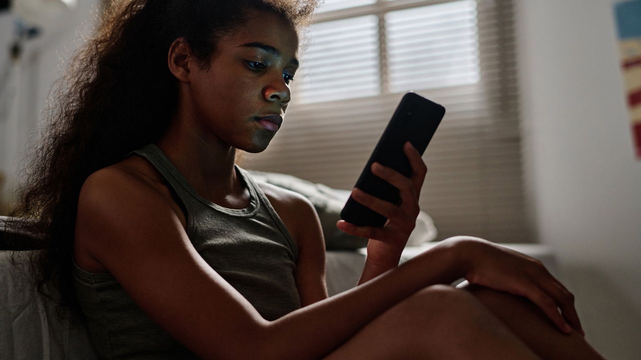 Side view of desperate teenager with cellphone sitting by bed in her room - stock photo