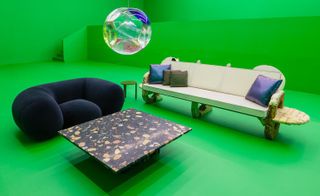 Lounge area with dark green velvet armchair, light coloured bench-style sofa and dark marble effect coffee table against a bright green wall and floor.