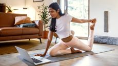 woman on an exercise mat doing a pilates move with a laptop in front of her in a living room setting 