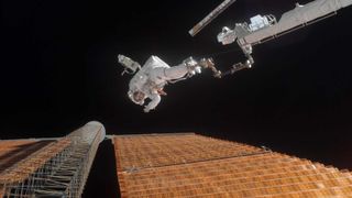 an astronaut on a robotic arm head-first over a solar panel, during a spacewalk