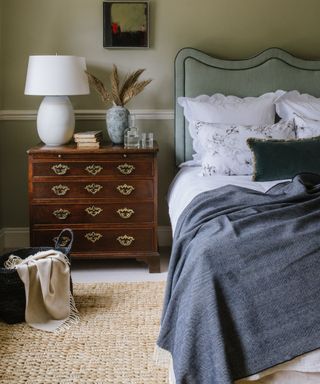 White bedding and blue blankets on a bed with an Anbôise headboard against a pale green wall beside a wooden dresser.