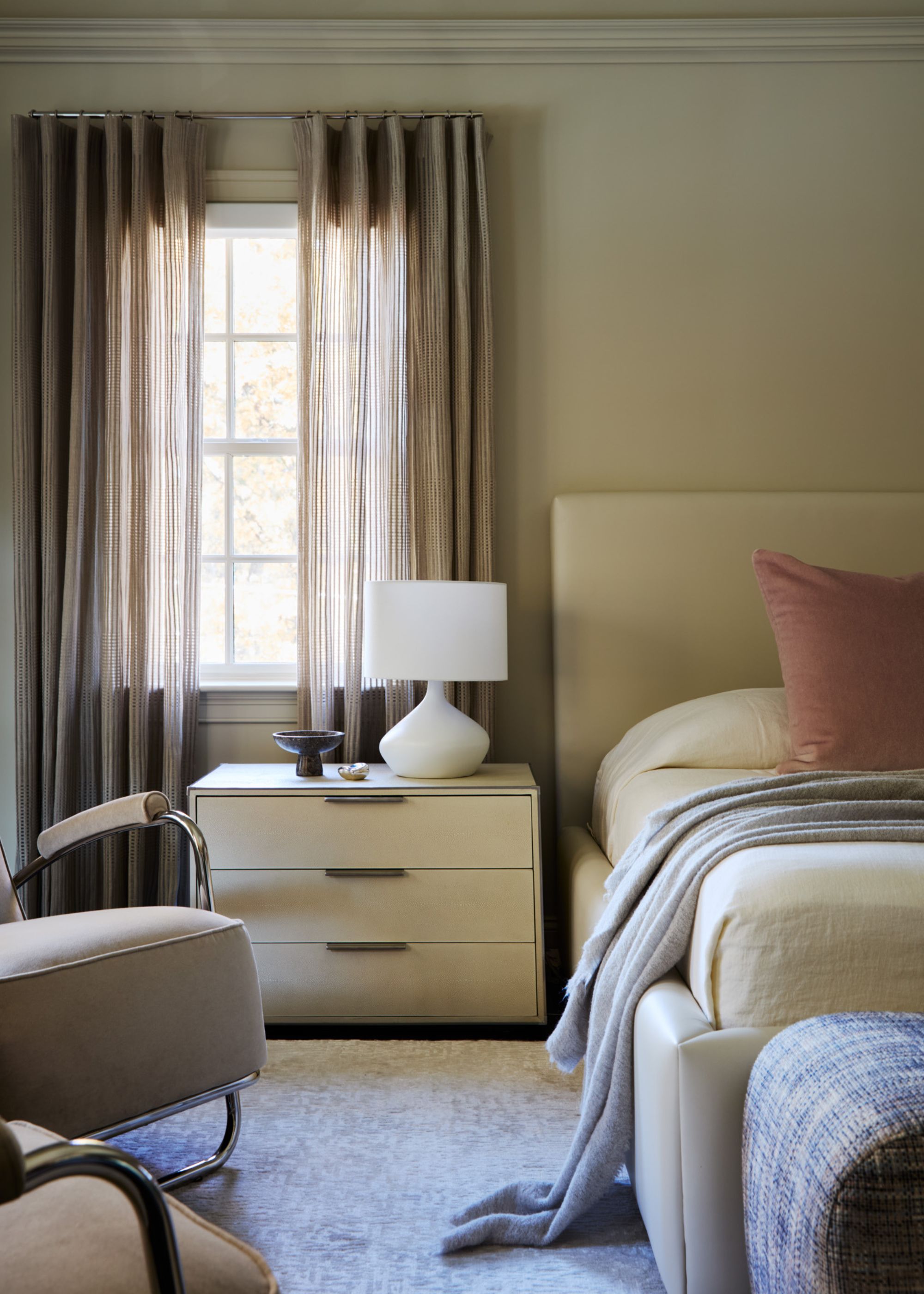 A bedroom with beige walls, a cream upholstered bed and taupe curtains.