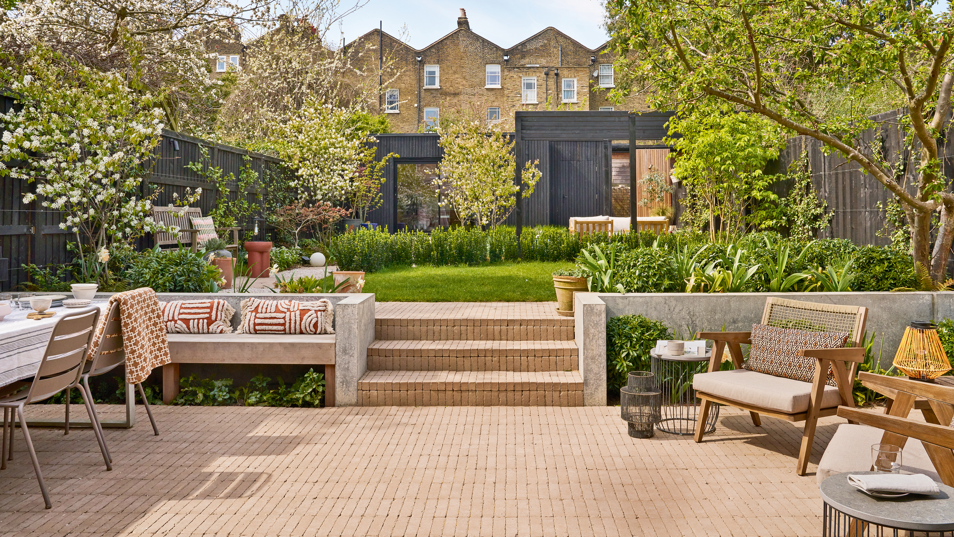 a large back garden with a black time framed studio at the end, trees and lawn in the middle and a brick patio in the foreground with a dining area on one side and a seating area on the other