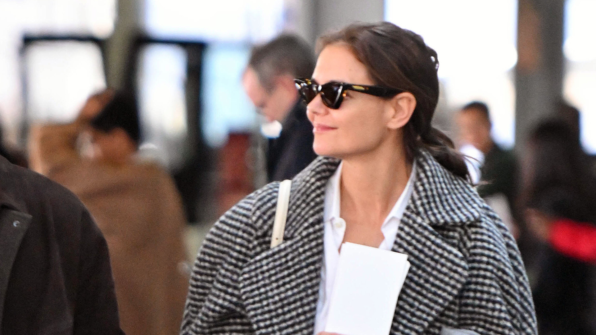 Katie Holmes at JFK airport wearing a printed black-and-white checked coat over a white button-down shirt with wide-leg jeans and black loafers