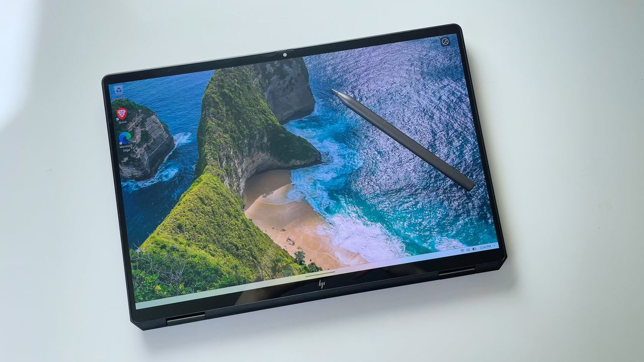 A top-down view of a laptop with its display folded back to make it look like a tablet; a digital pen is placed on top of the screen, which shows green hills leading to an ocean shore.