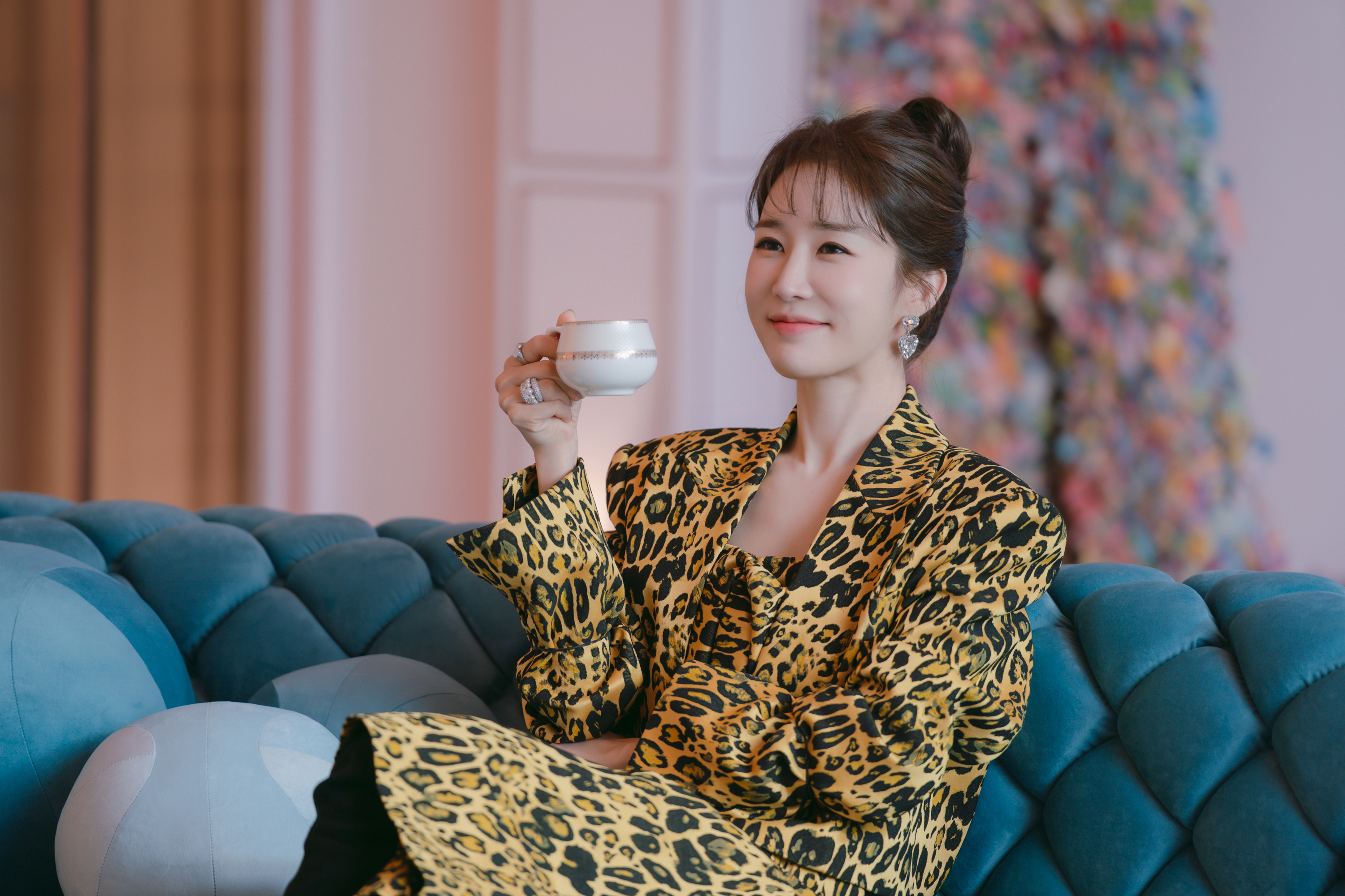 A woman (Yoo In-na as Dating Manager) wears a leopard-print coat while holding a teacup and sitting on a blue quilted couch in a pink room, in a still from the Netflix K-drama 'Boyfriend on Demand.'