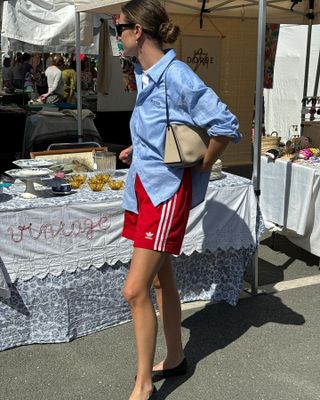 French woman wears red Adidas three stripe shorts, a blue button down and Chanel flats at an outdoor market.