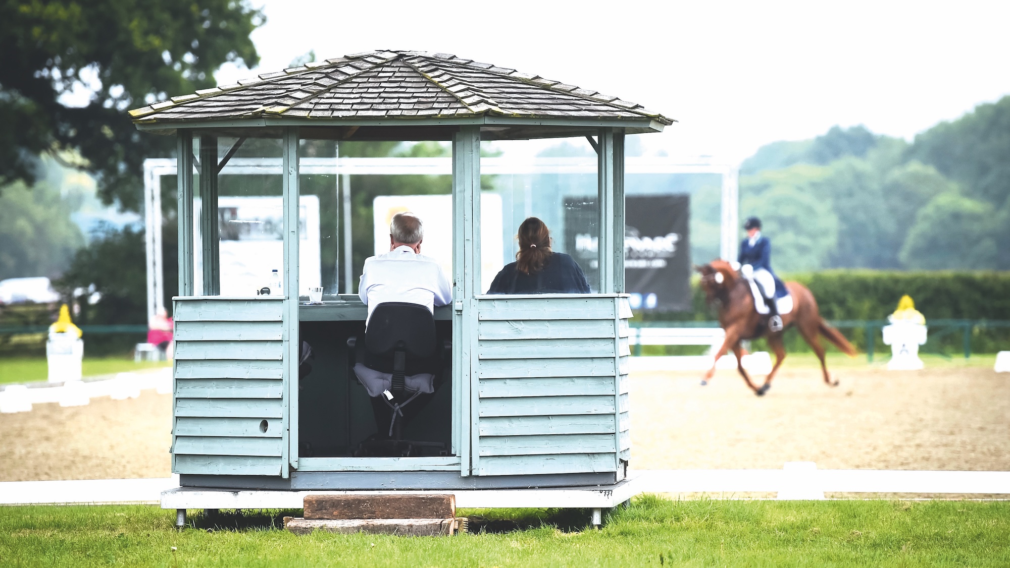 Image shows the back of a judges hut, and an unidentifiable chestnut horse and rider doing a dressage test