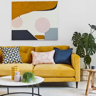 Mustard sofa with coloured cushions by artwork and houseplant
