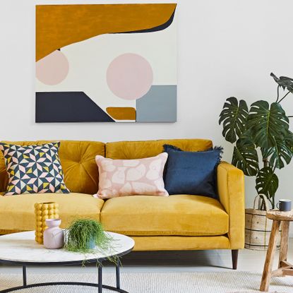 Mustard sofa with coloured cushions by artwork and houseplant