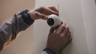 Installer in uniform puts security camera on wall fastening and connects it to system with cable. Man installs cameras in house. Concept of CCTV cameras, monitoring, safety and privacy.