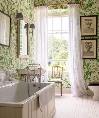 The 2021 bathroom trend for drapes shown in white alongside a green patterned wallpaper and panelled bath tub.