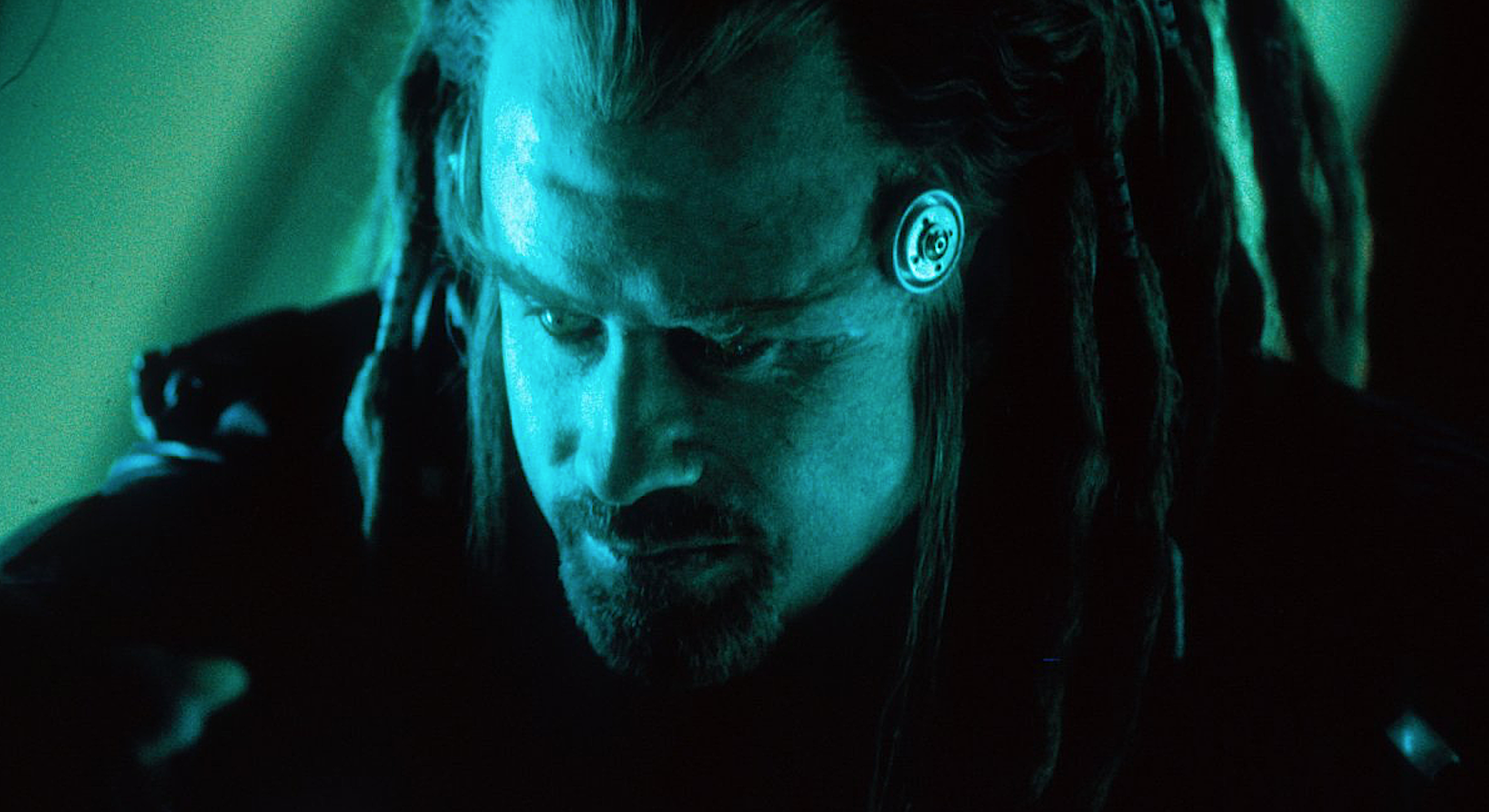 an alien male with dreadlock hair cast in a blue-green light