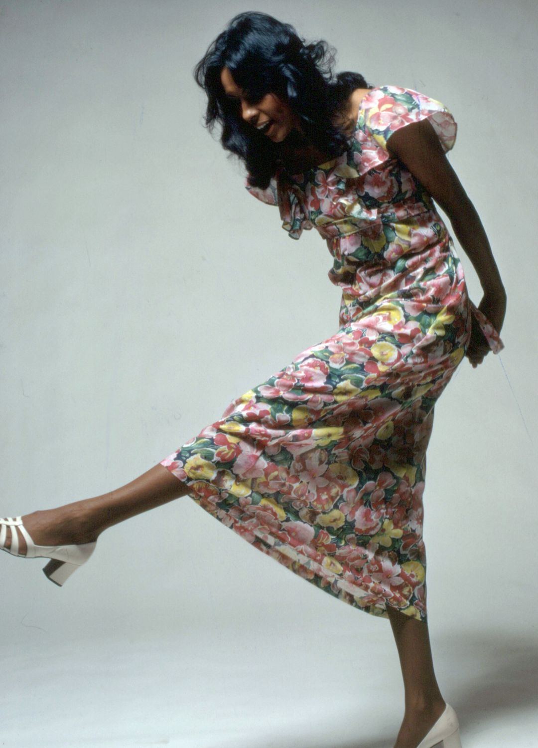 An unidentified woman models a summer dress with floral patterns and white chunky heeled shoes, New York, 1980s