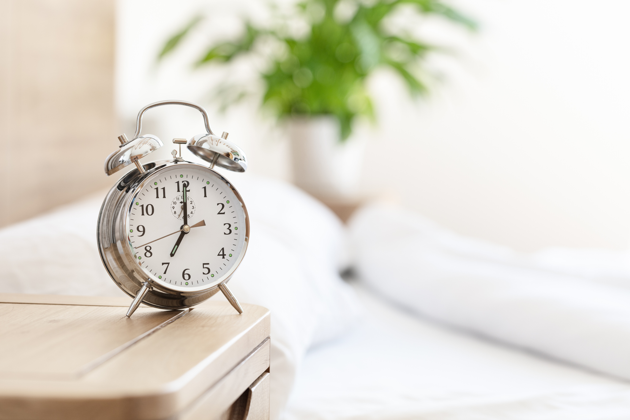 A metal alarm clock displaying seven o'clock in the morning in front of a light bedroom.