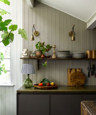 A small olive green kitchen with shiplap, two brass wall lights and a small table lamp