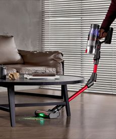 Arm holding Ultenic U16 Flex and vacuuming under dark coffee table in modern living room on brown, wooden floor.